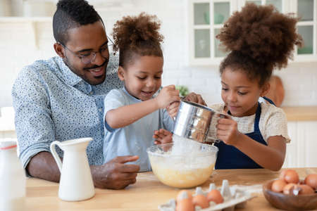 Caring african american dad cooking in kitchen with little ethnic kids make pastries together, happy...