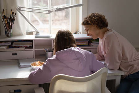 Smiling mother helping to teenage daughter with school assignment, sitting at work desk in bedroom,...
