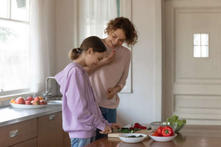 Happy mother and teenager daughter cooking salad, standing in modern kitchen at home together, smiling teen girl cutting fresh vegetables with knife, overjoyed mum and child preparing dinnerの写真素材