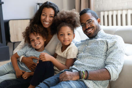 Portrait of smiling young african American family with two small kids sit relax on couch in living room together, happy biracial mom and dad hug cuddle with little children at home, show love careの写真素材