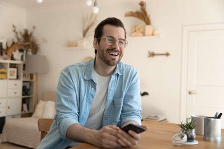 Happy millennial Caucasian guy in glasses sit at desk at home have fun talking chatting with friend,...