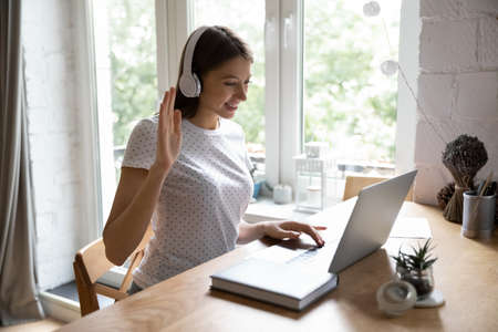 Smiling millennial girl in wireless headset sit at desk have online video call on laptop with teacher coach, happy young woman in headphones talk speak on webcam conference with friend or colleagueの写真素材