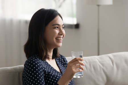 Smiling millennial Vietnamese girl relax on sofa at home drink clean clear mineral water from glass,...