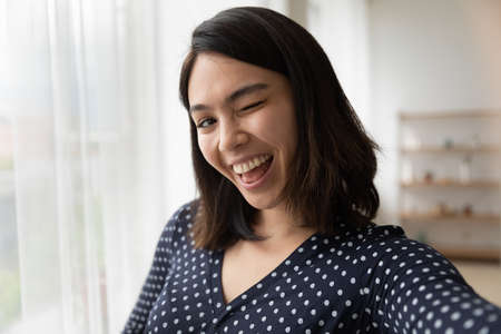 Self-portrait of funny happy millennial Asian girl have fun posing for photo on good quality smartphone camera at home, excited smiling Vietnamese young woman take selfie on modern cellphone gadgetの写真素材
