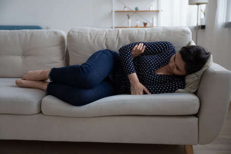 Sad young Vietnamese woman lying on sofa at home suffer after miscarriage or abortion, unhappy millennial Asian girl feel distressed upset struggle with depression or mental psychological problemsの写真素材