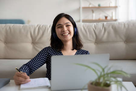 Overjoyed millennial Vietnamese girl in earphones have fun studying online on laptop at home, happy...