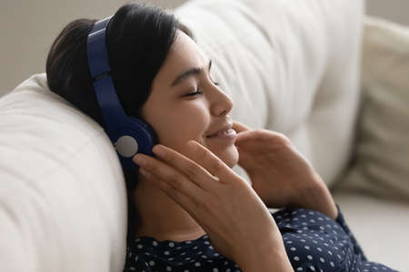 Close up of happy millennial Asian girl sit rest on couch listen to music in modern wireless...