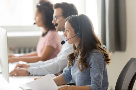 Call center agent in headset consulting client, woman in headphones working with documents, looking at computer screen, group employees sitting at workplace in customer support service officeの写真素材