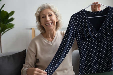 Elderly woman sit on couch show blouse look at camera, aged lady blogger share brand new wear,...