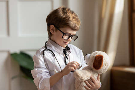 Smiling young kid boy in eyewear and white coat using tool, playing doctor patient game with...