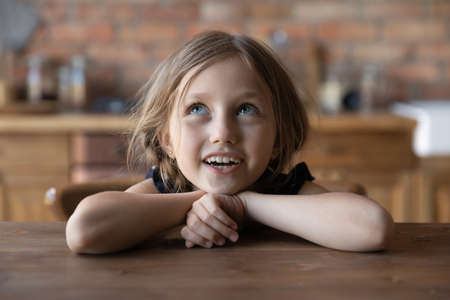Cute small preschooler blue-eyed girl with braids sit at table in kitchen have fun singing waiting for food, overjoyed little child kid relax at counter, enjoy happy childhood in house or homeの写真素材