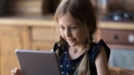 Happy little Caucasian girl look at tablet screen have fun play online application game on gadget,...