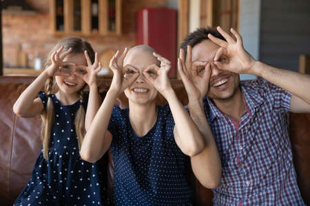 Portrait of overjoyed young family with little kid with sick cancer patient mother make funny faces relax at home together, happy man support ill hairless wife feel playful with small daughter childの写真素材