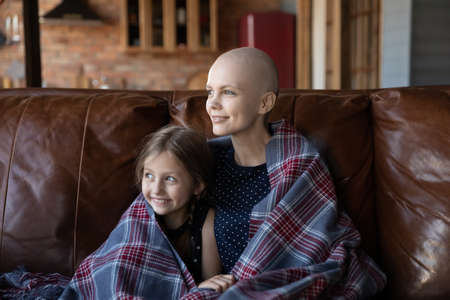 Happy young Caucasian sick cancer patient mom and little daughter sit relax on couch look in distance dream visualize, smiling hopeful ill hairless mother and small girl rest at home enjoy weekendの写真素材