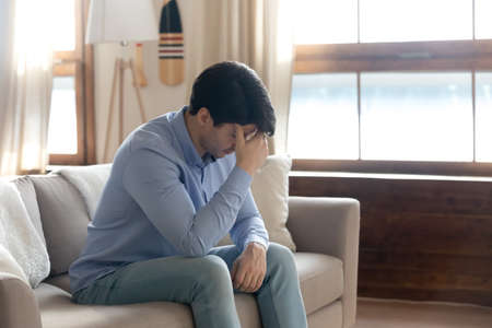 Side view stressed young guy touching forehead, feeling unwell alone in living room. Depressed millennial handsome man thinking of life problems, worrying about betrayal relations breakup at home.の写真素材