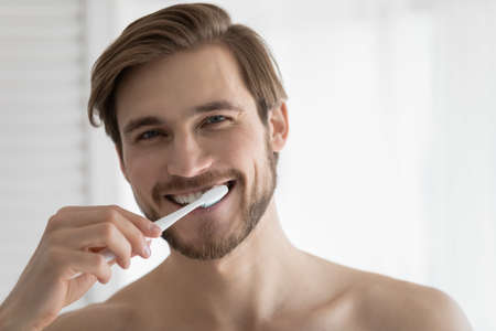 Head shot portrait close up smiling young man brushing teeth, looking at camera, personal oral hygiene concept, satisfied handsome guy enjoying morning routine, standing in bathroomの写真素材