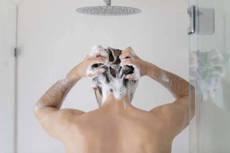 Rear view young man cleaning hair with foamy anti dandruff shampoo close up, standing in modern bathroom at home, taking morning shower, washing head and body, personal hygiene and skincare conceptの写真素材