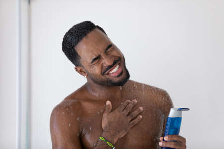 Close up smiling overjoyed African American handsome young man taking shower, holding shampoo or moisturizing gel bottle, relaxing, standing in bathroom, enjoying morning routine procedureの写真素材