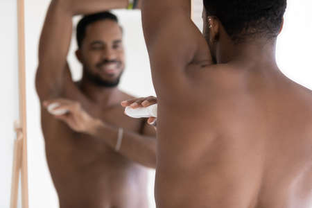 Rear view smiling African American handsome young man applying deodorant on armpit, standing in bathroom, looking in mirror, enjoying morning routine procedure, using stick underarm deodorantの写真素材