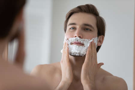 Mirror reflection head shot close up young man applying shaving foam, standing in bathroom, handsome satisfied male doing personal hygiene, skincare and morning routine procedure at homeの写真素材