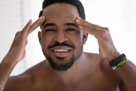 Head shot portrait smiling African American young man with naked shoulders checking wrinkles or acne on forehead, looking at camera, facial cosmetology, treatment, skincare conceptの写真素材