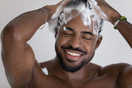 Close up smiling African American handsome young man washing cleaning hair with foamy anti-dandruff shampoo, taking shower, enjoying morning routine procedure, standing in bathroomの写真素材