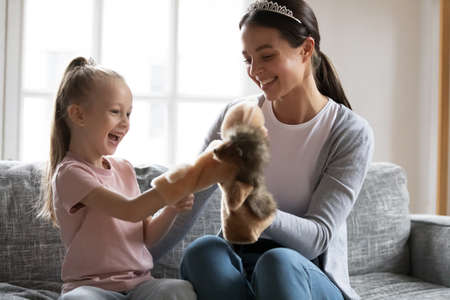 Overjoyed little adorable preschool blond girl playing hand toys with loving young mother, spending leisure time together indoors, relaxing on sofa. happy different generations family pastime concept.の写真素材