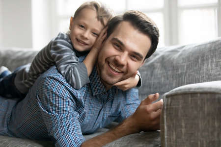 Head shot portrait of adorable little son lying on fathers back, resting together on comfortable sofa. Smiling two generations family enjoying sweet relaxed weekend free time moment at home.の写真素材