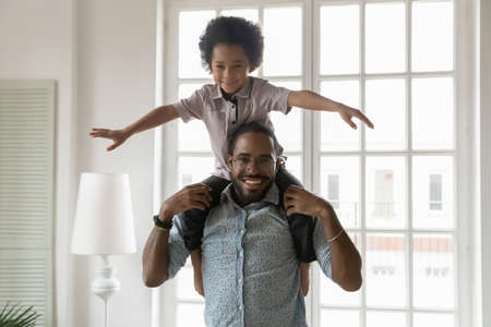 Portrait of little ethnic boy child sit on shoulders play with excited african American dad at home, smiling young biracial father carry on back, have fun enjoy family weekend together with small sonの写真素材