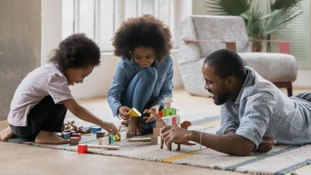 Loving young african American father sit on floor in living room play with toys blocks with small ethnic children, playful biracial father enjoy constructing with wooden bricks with little kidsの写真素材
