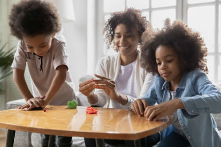 Excited african American mom or nanny have fun engaged in creative activity with little kids, happy biracial mother play with plasticine playdough, enjoy weekend with small children, hobby conceptの写真素材