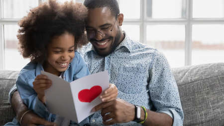 Happy young biracial father and small ethnic daughter sit on couch at home reading handmade postcard together, loving little african American girl present card greeting with birthday smiling dadの写真素材