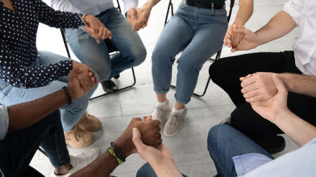 Close up diverse patients sitting on chairs in circle, holding hands, expressing support and unity...