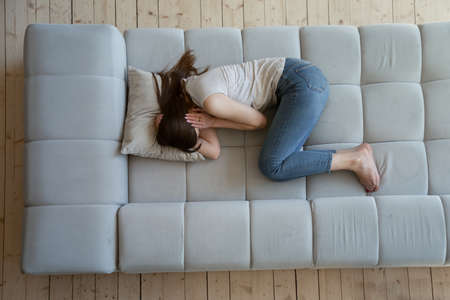 Distressed young female student lying on couch snuggling down and hugging head having panic or...