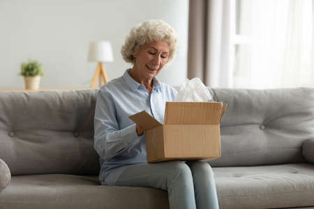 Positive middle aged older woman sitting on couch with carton box on laps, feeling curious about...