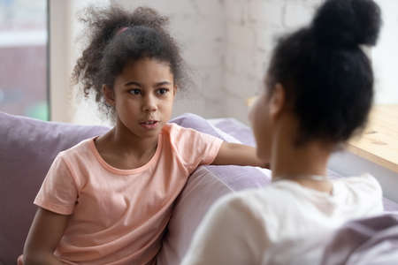 Sharing secrets with mom. Preteen african girl sitting on couch having confident frank talk with understanding mother or elder sister, school psychologist listening to bullying or racial abuse victimの写真素材