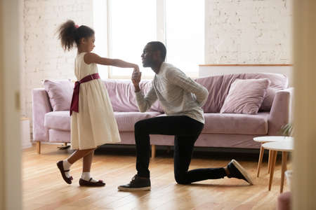 May I have this dance, my princess. Caring african father standing on one knee engaging gentle his little daughter for a waltz. Black preteen girl in ball gown dancing in living room with beloved dadの写真素材