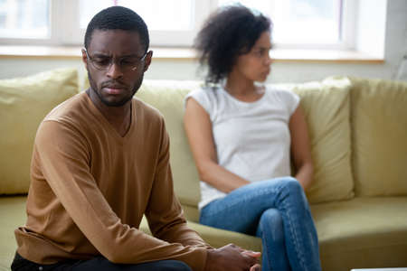Family argument. Millennial african married couple, spouses or lovers sitting on coach separating after quarrel or breaking up. Sad angry black pair husband and wife resenting and ignoring each otherの写真素材