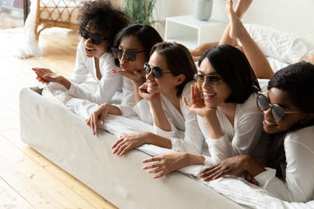 Funny diverse girls wearing stylish trendy sunglasses equal bathrobes lying in bed posing photographing in hotel bedroom. After spa procedures five women enjoy photoshoot celebrating hen-party conceptの写真素材