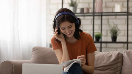 Smiling millennial Caucasian girl in headphones sit on sofa in living room study online on laptop...