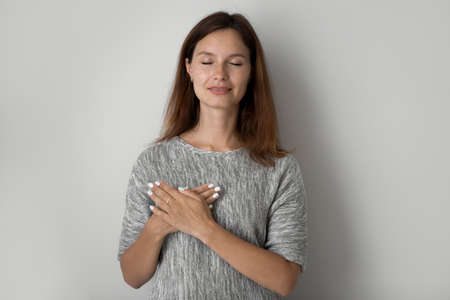 Hopeful young Caucasian woman isolated on grey studio background hold keep hands on heart chest feel...