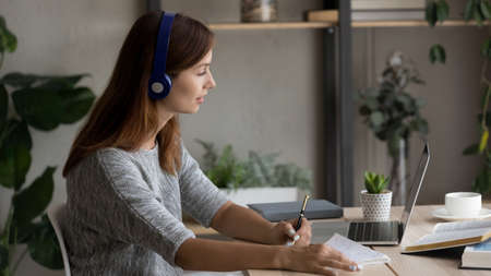Young Caucasian woman in headphones sit at desk work look at laptop screen take online course,...