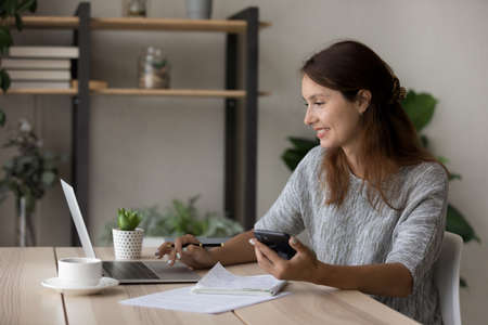 Happy young Caucasian woman sit at desk look at laptop screen use cellphone study online from home,...
