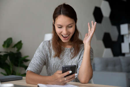 Excited millennial Caucasian female look at smartphone screen triumph get pleasant good text...