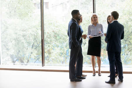 Diverse group of business people stand relaxedly in office corridor during coffee break, consulting,...