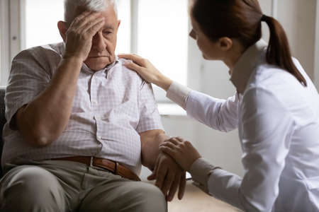 Unhealthy mature old patient complaining to doctor about head ache migraine illness at checkup meeting in clinic. Professional young female general practitioner giving advice, treating elderly man.の写真素材