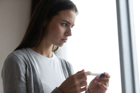 Close up anxious unhappy woman holding plastic pregnancy test kit in hands, stressed frustrated young female dissatisfied by result, health problem, infertility, unwanted pregnancy, standing at homeの写真素材