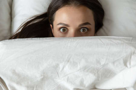 Close up top view funny young woman peeking from duvet, scared shocked female looking at camera,...