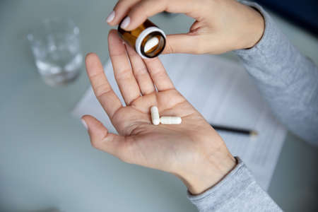 Close up young woman taking out pills from packaging, sitting at table with glass of water,...