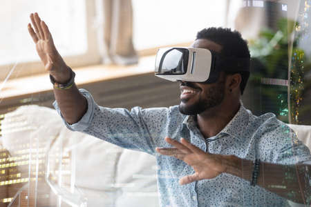 Smiling African American man in VR glasses enjoying augmented reality close up, sitting on couch at...